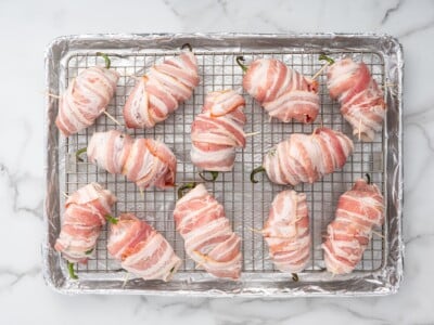 Twelve bacon-wrapped jalapeños secured with toothpicks on a wire rack over a foil-lined baking sheet, ready to go in the oven.