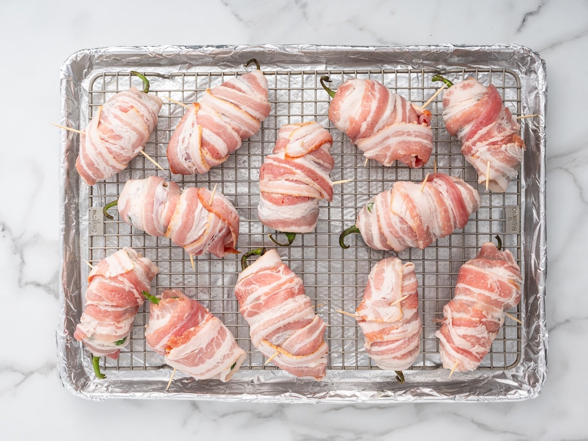 Twelve bacon-wrapped jalapeños secured with toothpicks on a wire rack over a foil-lined baking sheet, ready to go in the oven.