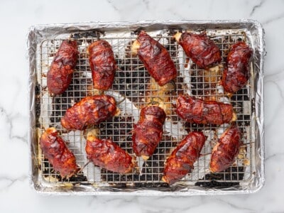 Twelve fully baked Texas Twinkies on a wire rack over a foil-lined baking sheet, glazed with caramelized BBQ sauce and crispy bacon.