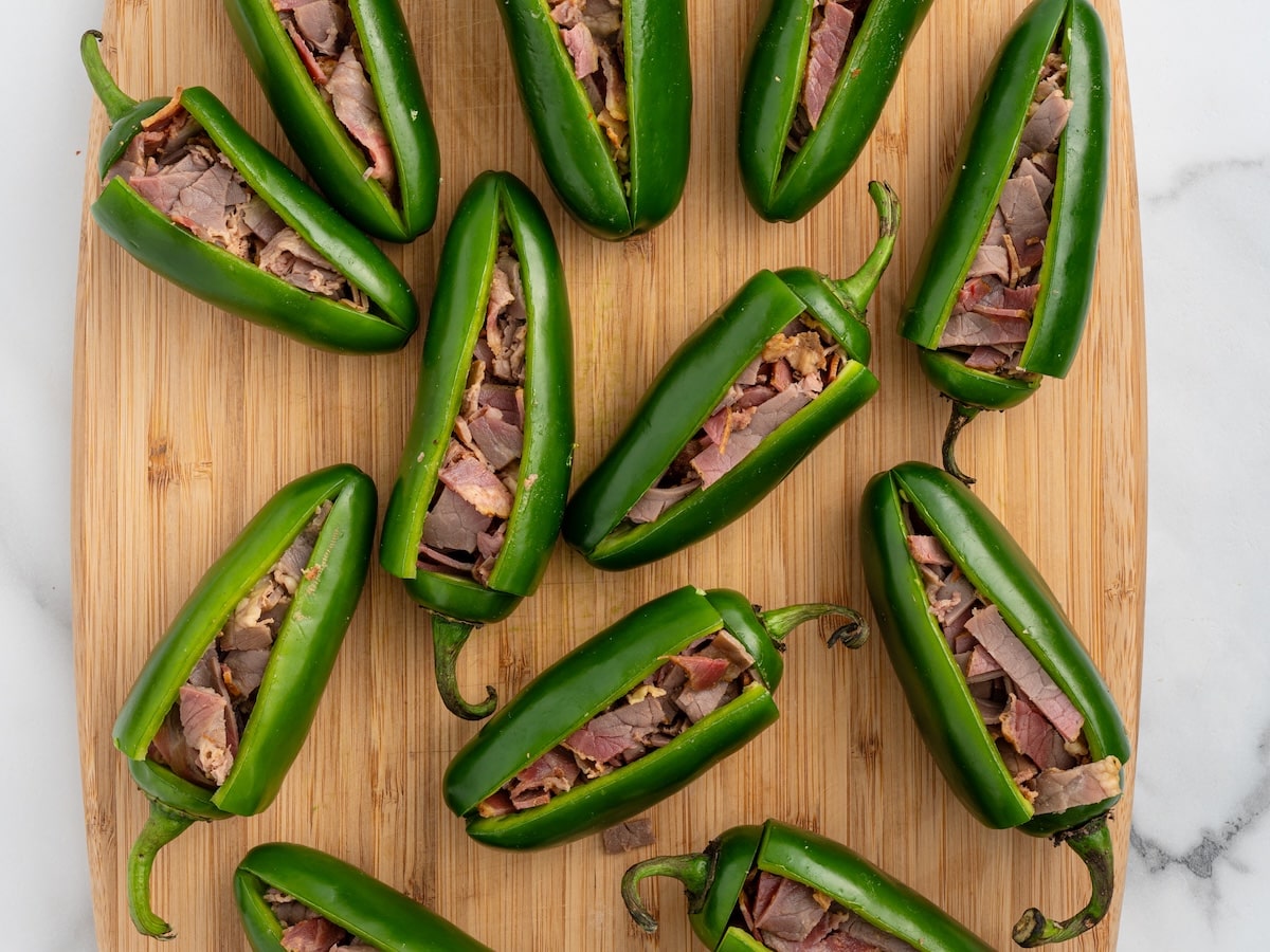 Twelve jalapeños sliced open and stuffed with chopped smoked beef brisket, arranged on a wooden cutting board.