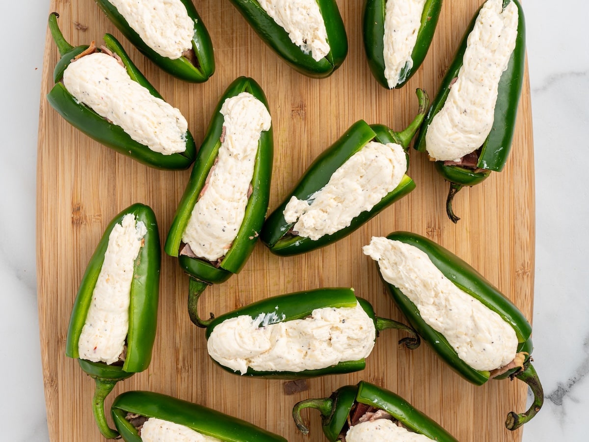 Jalapeños filled with chopped brisket and topped with a generous layer of seasoned cream cheese mixture, arranged on a wooden cutting board.