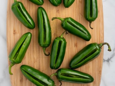 Twelve fresh jalapeños with T-shaped cuts sliced into each one, arranged on a wooden cutting board ready to be stuffed.