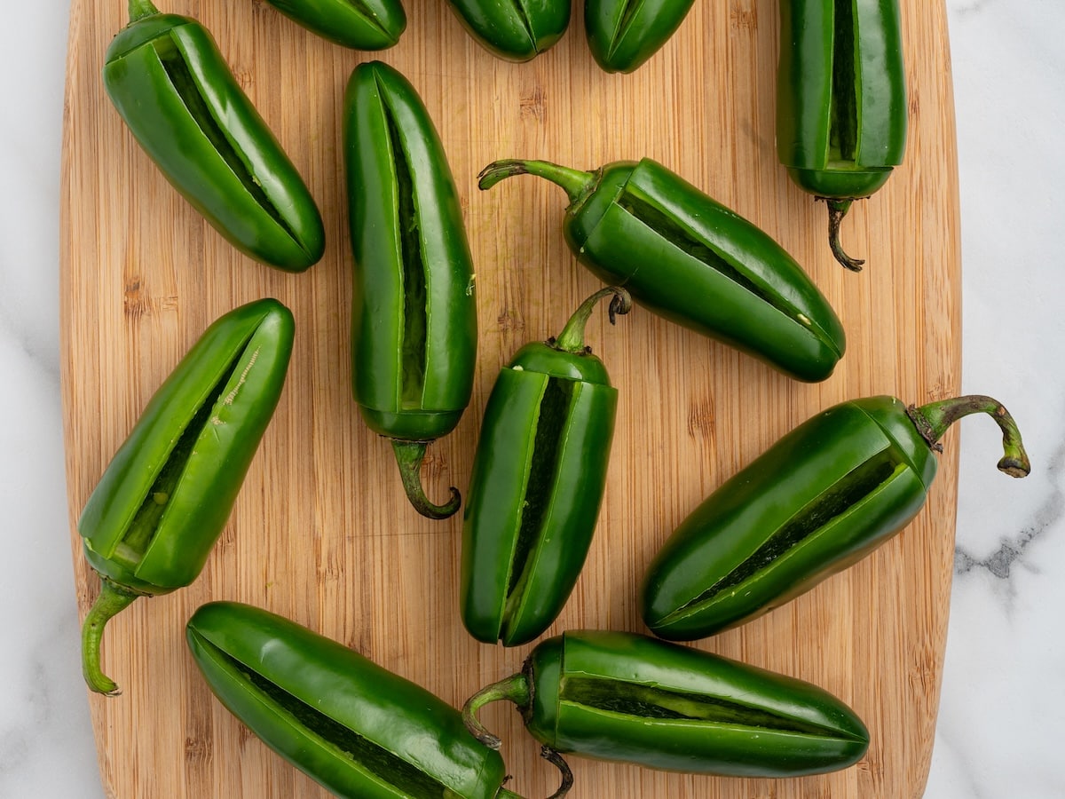 Twelve fresh jalapeños with T-shaped cuts sliced into each one, arranged on a wooden cutting board ready to be stuffed.