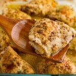 Close-up of a baked chicken thigh lifted from the baking dish with a wooden spoon, showcasing the juicy interior and flavorful lemon garlic pan juices. This easy baked chicken thighs recipe features bone-in chicken seasoned with Dijon mustard, garlic, lemon, and Italian seasoning.