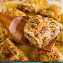 Close-up of a baked chicken thigh lifted from the baking dish with a wooden spoon, showcasing the juicy interior and flavorful lemon garlic pan juices. This easy baked chicken thighs recipe features bone-in chicken seasoned with Dijon mustard, garlic, lemon, and Italian seasoning.