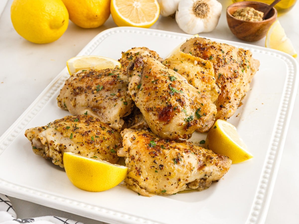 Juicy baked chicken thighs arranged on a white serving platter and garnished with fresh lemon wedges. These bone-in chicken thighs are seasoned with garlic, Dijon mustard, Italian herbs, and lemon, creating an easy oven-baked chicken dinner with crispy golden skin and tender meat.