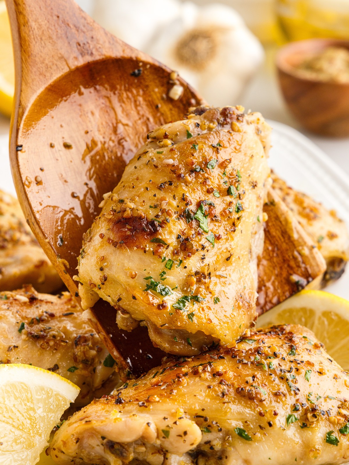 Close-up of juicy baked chicken thighs being lifted with a wooden spoon, highlighting the golden skin coated in a lemon garlic Dijon sauce with visible herbs and seasonings. Fresh lemon wedges frame this easy baked chicken thighs recipe, perfect for a flavorful weeknight dinner.