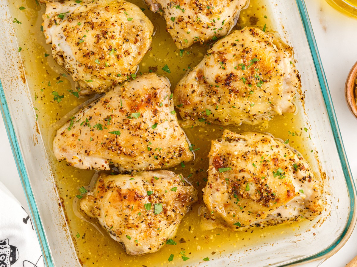 Overhead view of baked chicken thighs resting in a glass baking dish filled with buttery lemon garlic pan juices. The bone-in chicken thighs are seasoned with Dijon mustard, Italian herbs, garlic, and lemon, then roasted until tender and lightly golden for an easy family dinner recipe.