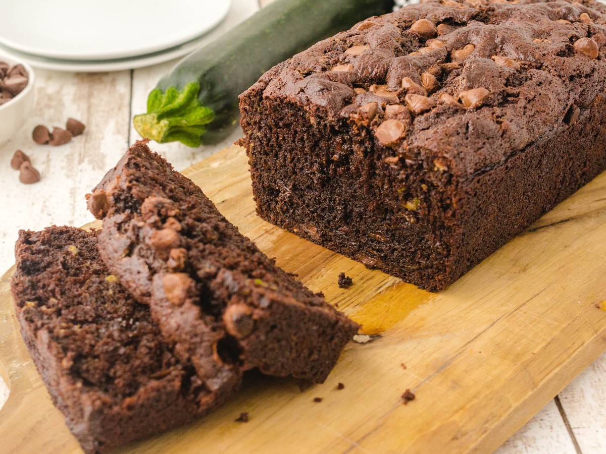 Loaf of Chocolate Zucchini Bread with sliced pieces on a wooden board, chocolate chips, and zucchini in the background.