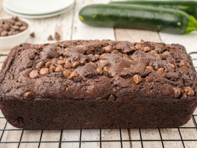 Freshly baked loaf of Chocolate Zucchini Bread studded with chocolate chips cooling on a wire rack with whole zucchini in the background.