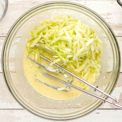 Overhead photo of shredded zucchini being added to the wet batter for Chocolate Zucchini Bread with electric mixer beaters resting in the bowl.