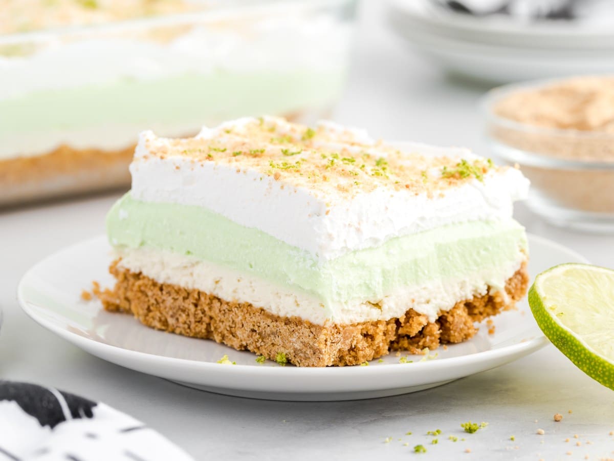 Slice of Key Lime Pie Lasagna on a white plate with graham cracker crust, creamy layers, whipped topping, lime zest, and a fresh lime half.