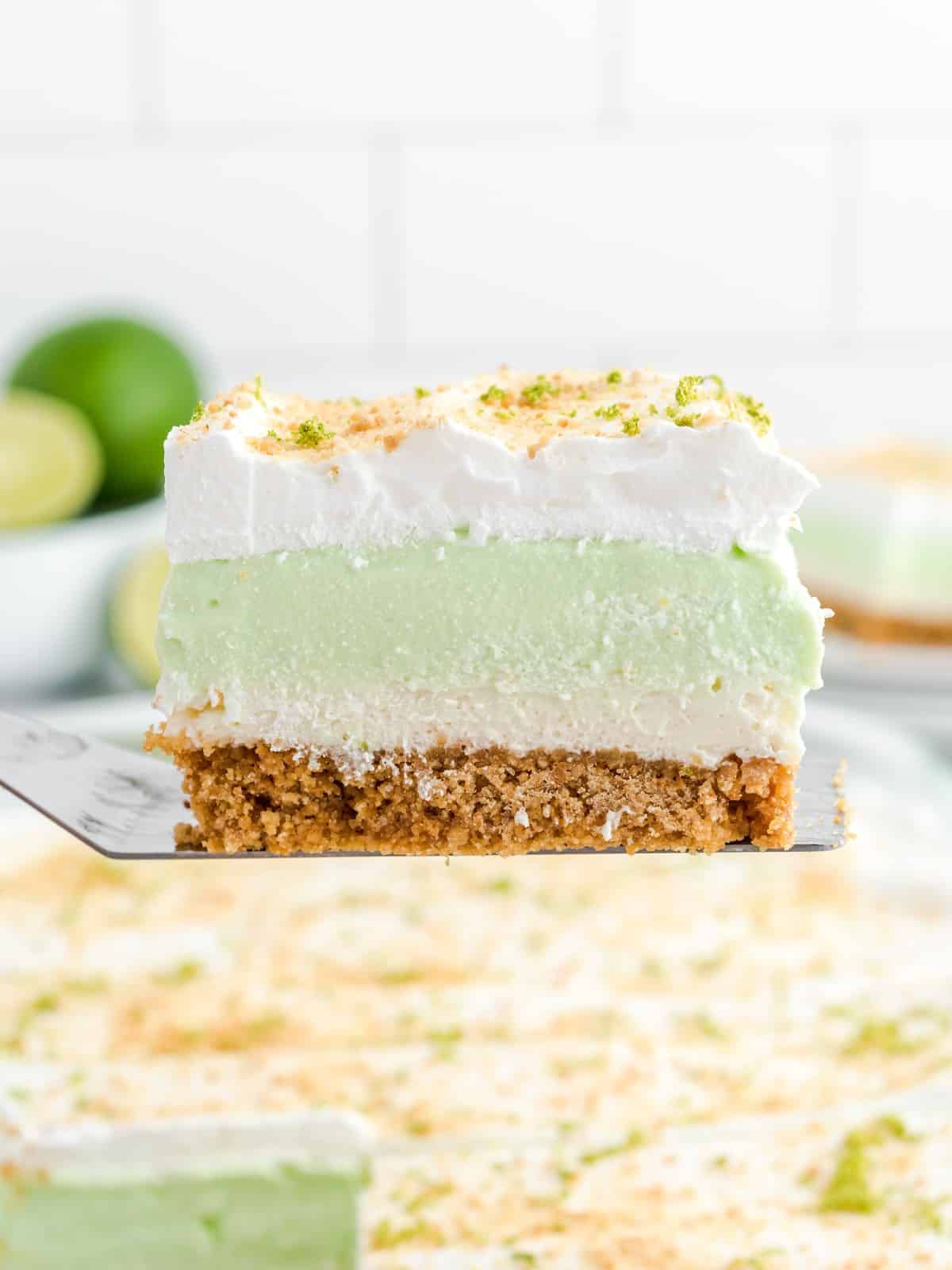 Slice of Key Lime Pie Lasagna lifted on a spatula, showing distinct layers of graham cracker crust, cream cheese filling, creamy key lime layer, and whipped topping sprinkled with lime zest and graham cracker crumbs.