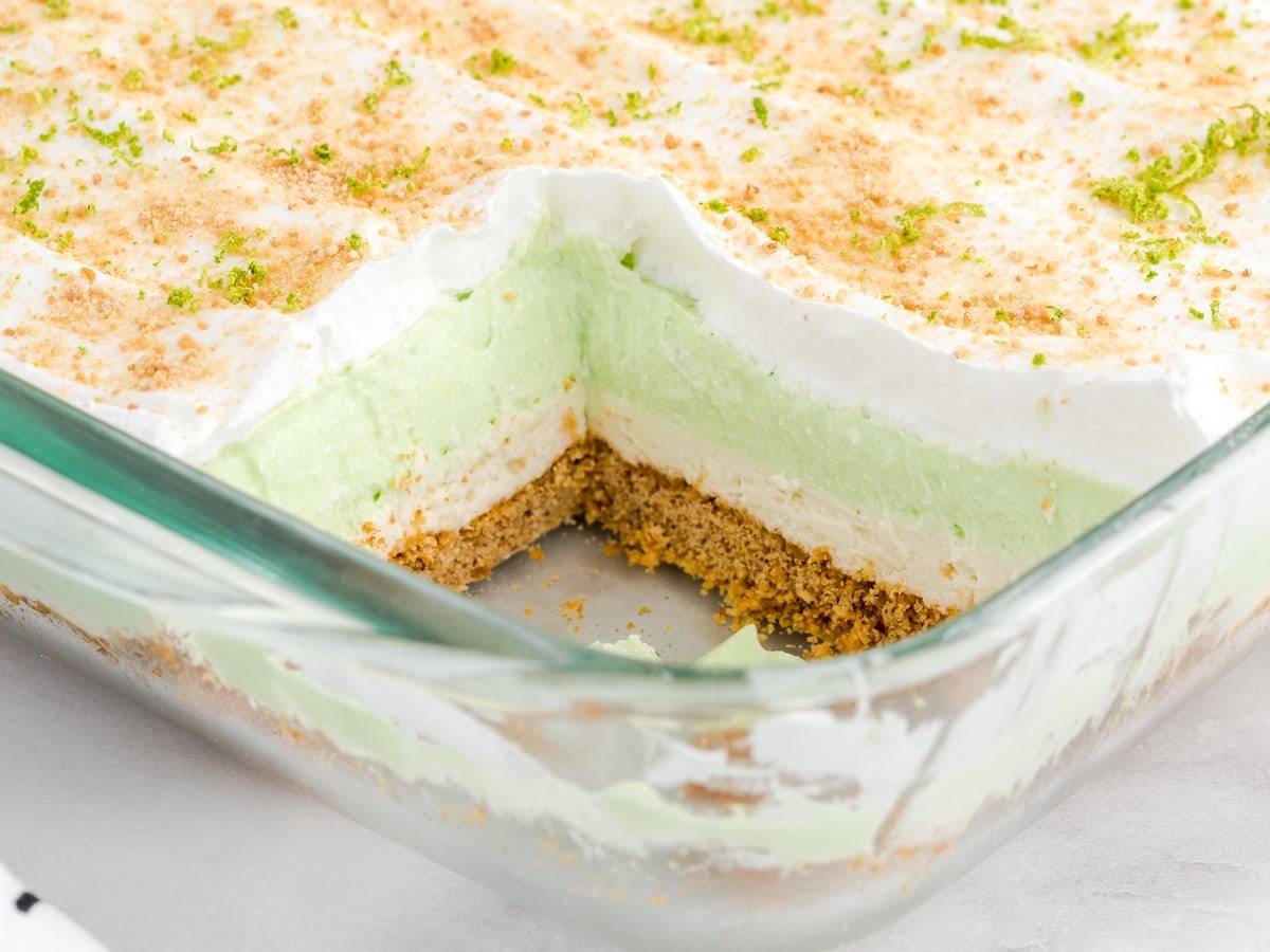 Key Lime Pie Lasagna in a glass baking dish with a slice removed, revealing layers of graham cracker crust, cream cheese filling, smooth key lime pudding layer, and whipped topping garnished with lime zest and graham cracker crumbs.
