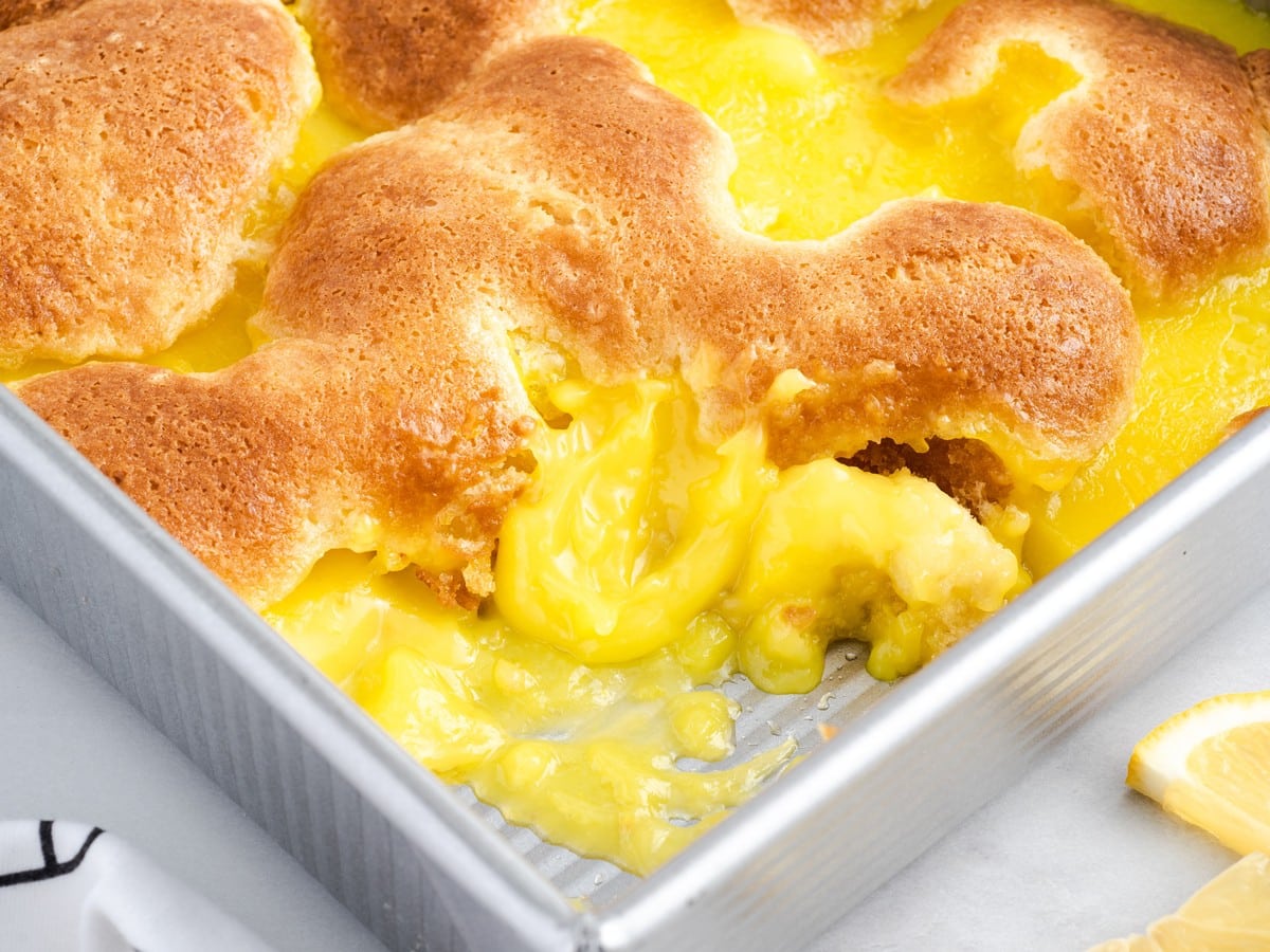 Freshly baked Lemon Cobbler in a baking dish with golden cobbler topping and rich lemon filling bubbling through the cracks, showing a scooped serving removed from the corner.