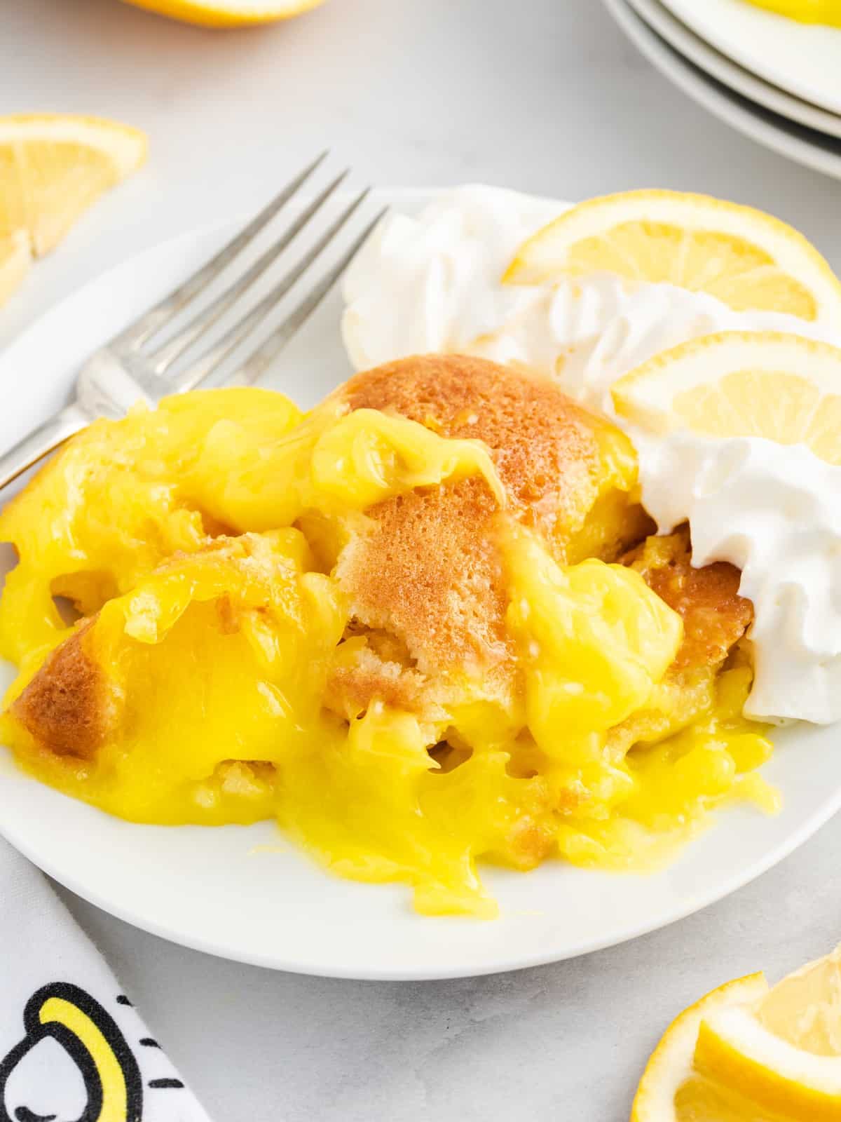 Individual serving of warm Lemon Cobbler on a white dessert plate topped with creamy lemon filling, golden cobbler crust, whipped cream, and fresh lemon slices.