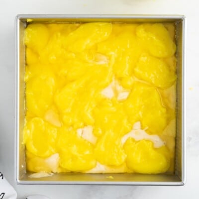 Lemon pie filling spooned evenly over the batter and melted butter in a square baking dish, creating the signature layered base for homemade Lemon Cobbler before baking.