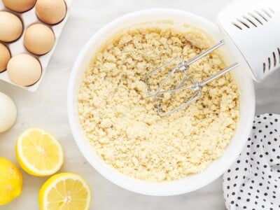 Crumbly cake mix dough in a white mixing bowl with a hand mixer, surrounded by fresh lemons and eggs on a marble surface.