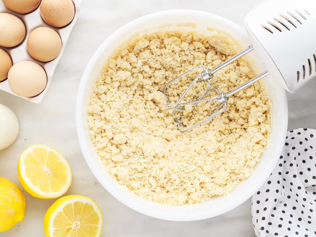 Crumbly cake mix dough in a white mixing bowl with a hand mixer, surrounded by fresh lemons and eggs on a marble surface.
