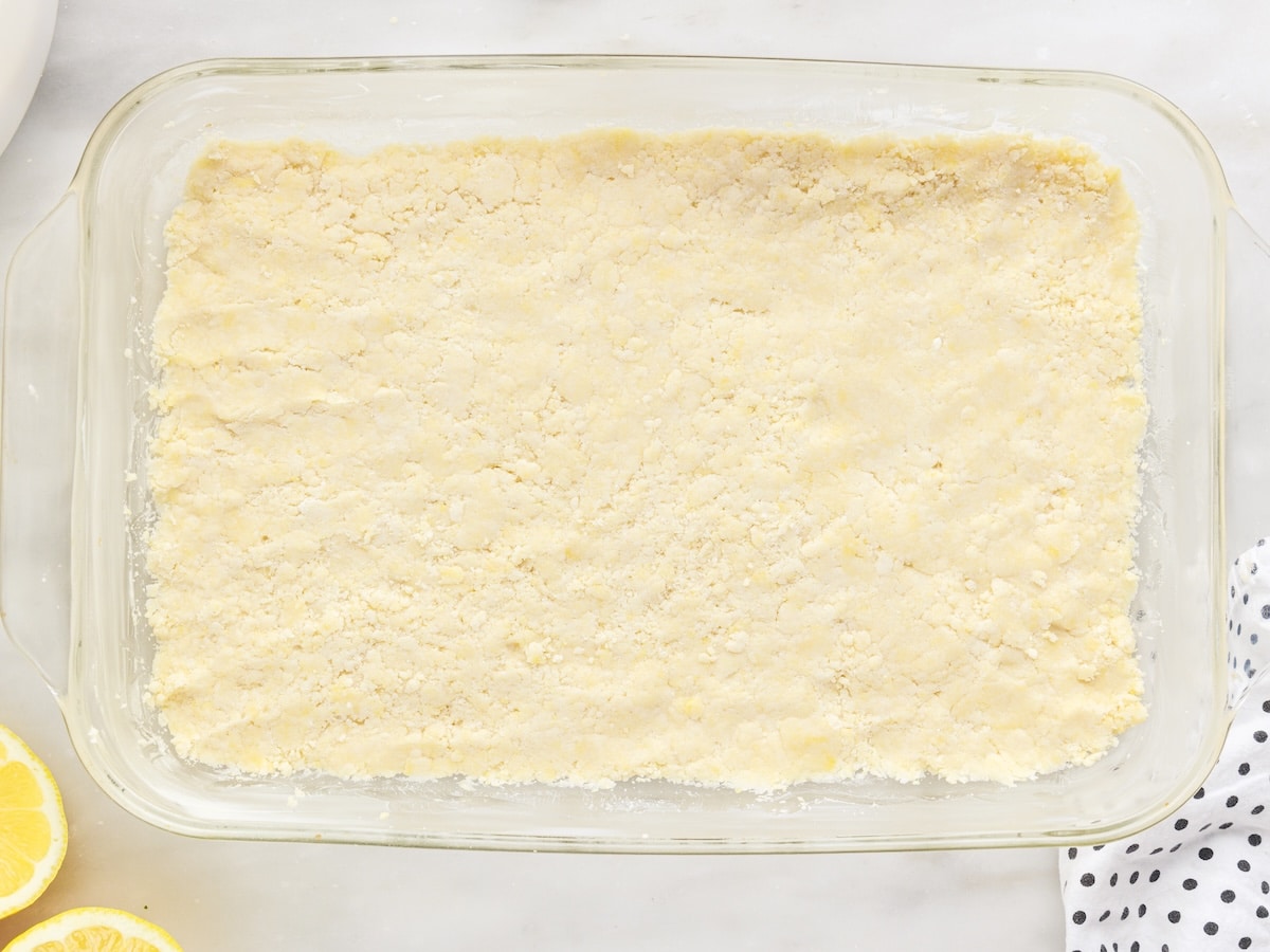 Par-baked cake mix crust pressed into a glass 9x13 baking dish, with sliced lemons visible alongside.