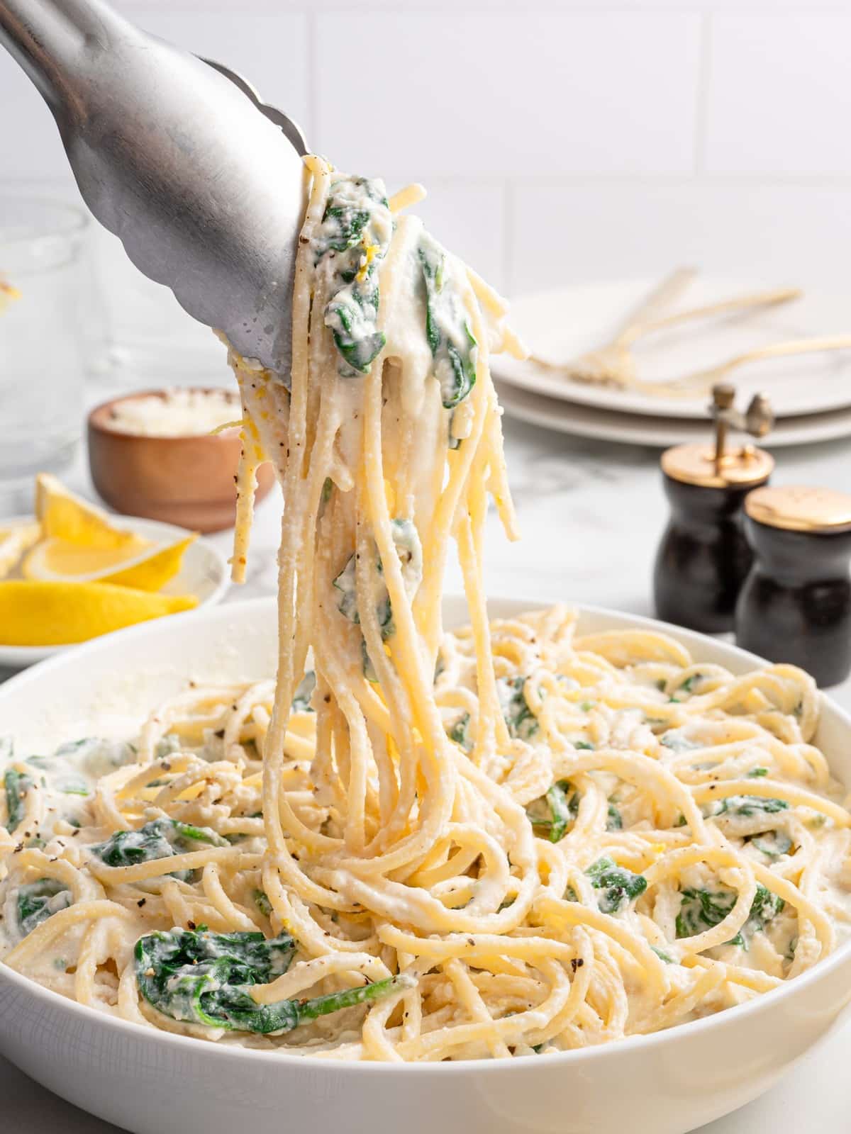 Creamy Lemon Ricotta Pasta with spinach being lifted from a white serving bowl with tongs, showing spaghetti coated in ricotta parmesan sauce with lemon zest and black pepper.