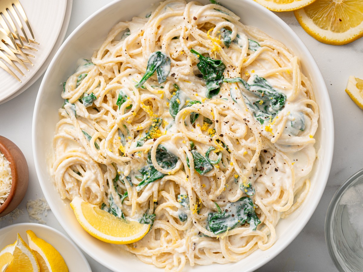 Overhead view of creamy Lemon Ricotta Pasta made with spaghetti and baby spinach in a rich ricotta parmesan sauce, garnished with lemon zest, cracked black pepper, and fresh lemon wedges.