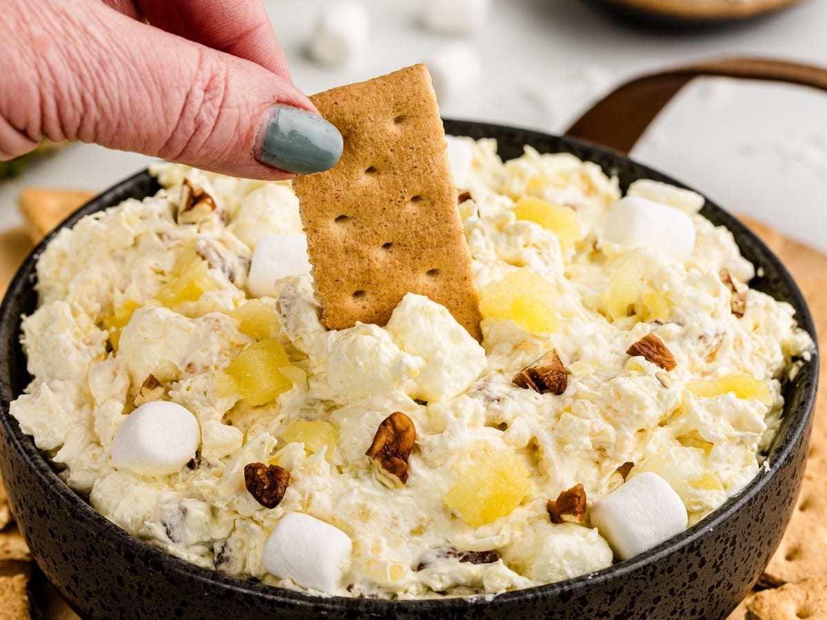 Pineapple Fluff in a black bowl topped with mini marshmallows, pineapple pieces, and chopped pecans, with a graham cracker being dipped into the creamy dessert.