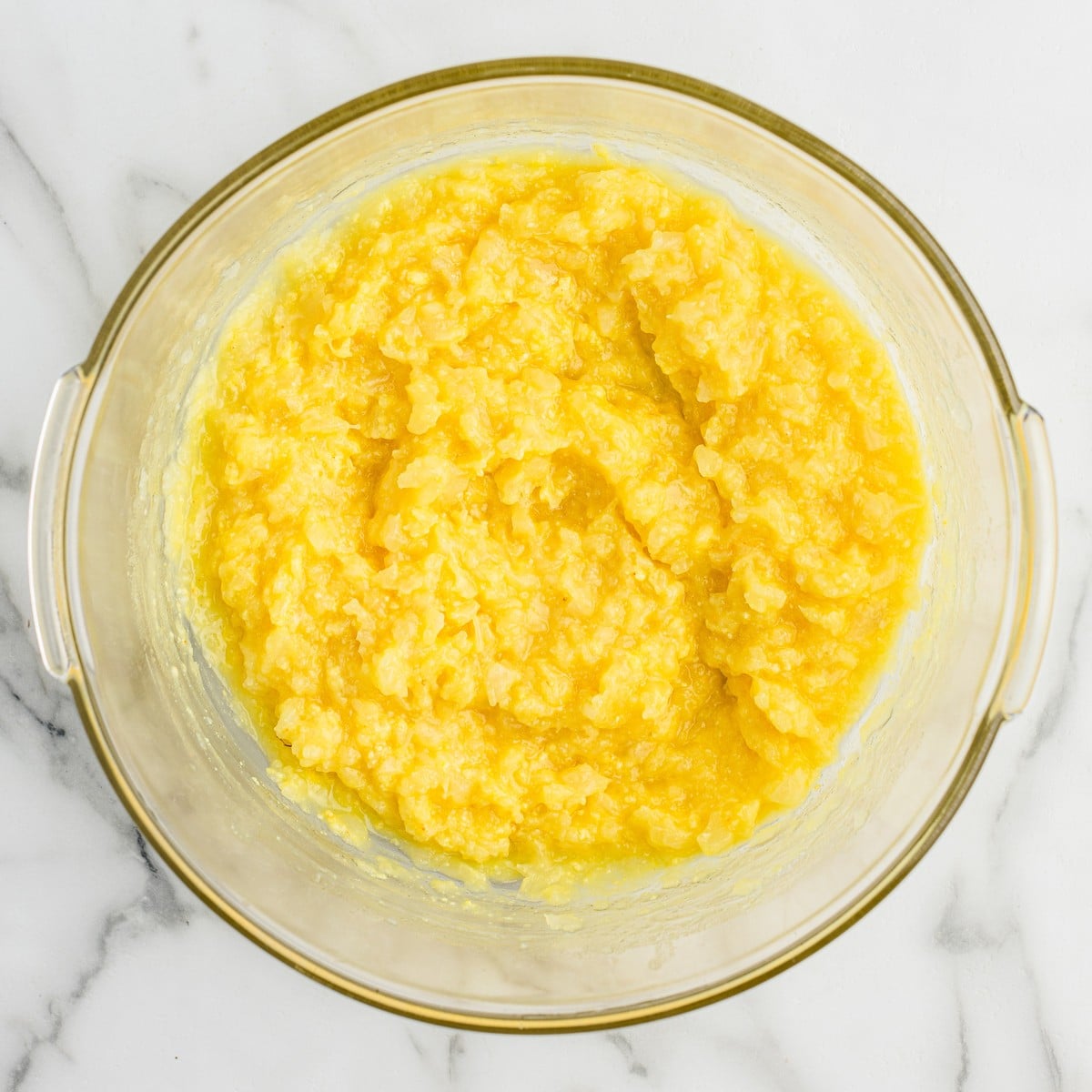 Overhead photo of crushed pineapple mixed with vanilla instant pudding in a glass bowl, creating the creamy yellow base for Pineapple Fluff dessert salad.