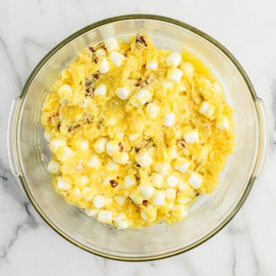 Overhead view of Pineapple Fluff ingredients combined in a glass mixing bowl, showing crushed pineapple mixture mixed with mini marshmallows and chopped pecans before adding whipped topping.