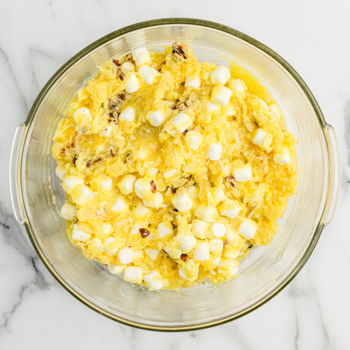 Overhead view of Pineapple Fluff ingredients combined in a glass mixing bowl, showing crushed pineapple mixture mixed with mini marshmallows and chopped pecans before adding whipped topping.