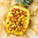 Overhead view of fresh pineapple salsa served inside a hollowed pineapple half, filled with diced pineapple, tomatoes, red onion, and herbs. The pineapple bowl sits on a white platter surrounded by tortilla chips, creating a colorful and tropical party appetizer presentation.