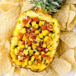 Overhead view of fresh pineapple salsa served inside a hollowed pineapple half, filled with diced pineapple, tomatoes, red onion, and herbs. The pineapple bowl sits on a white platter surrounded by tortilla chips, creating a colorful and tropical party appetizer presentation.