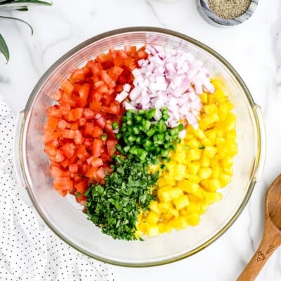 Overhead view of a glass bowl filled with freshly chopped pineapple salsa ingredients, including diced pineapple, tomatoes, red onion, jalapeño, and cilantro arranged separately before mixing.