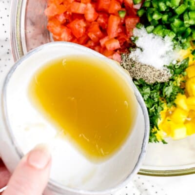 Close-up of fresh pineapple salsa ingredients in a glass bowl with lime juice being poured over diced pineapple, tomatoes, jalapeños, cilantro, salt, and black pepper before mixing.