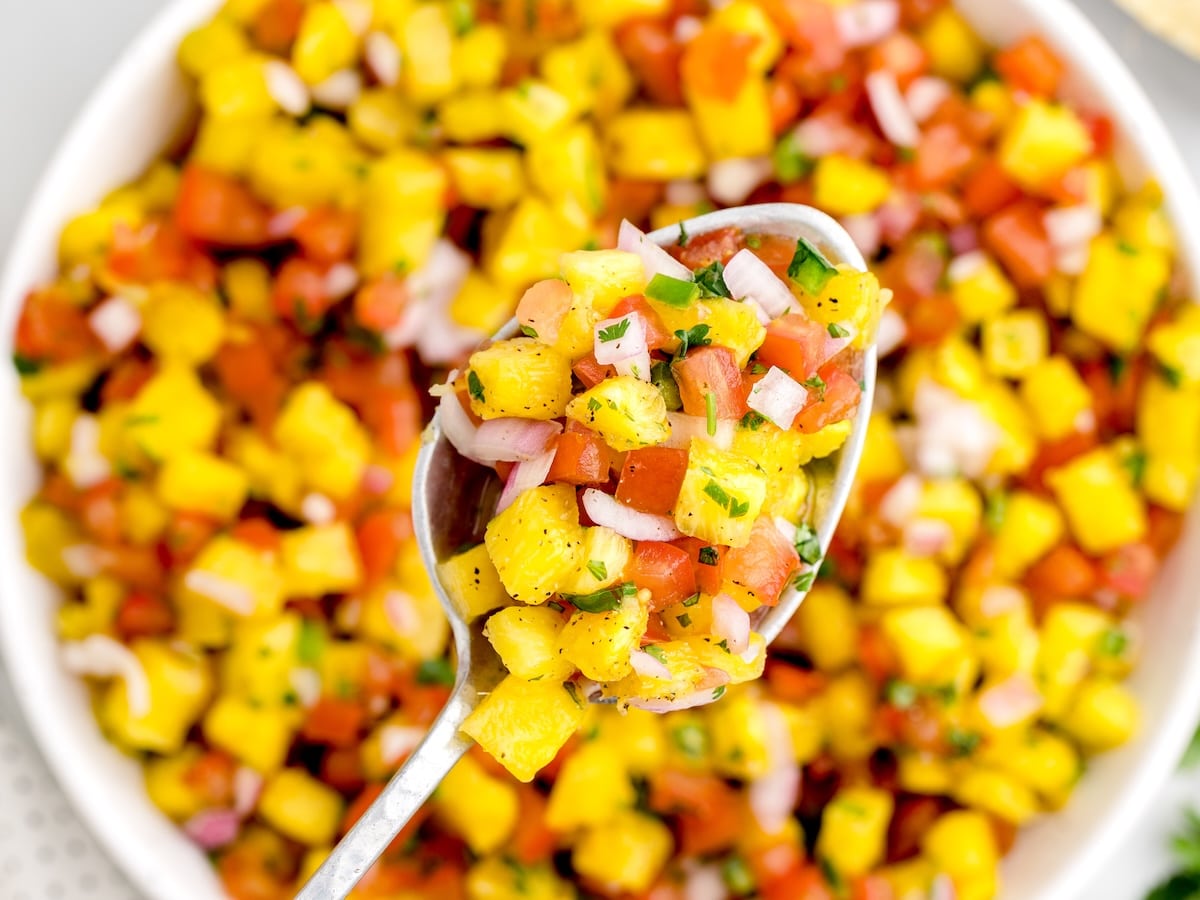 Close-up of a spoonful of fresh pineapple salsa with diced pineapple, tomatoes, red onion, and cilantro held over a bowl of colorful homemade salsa.