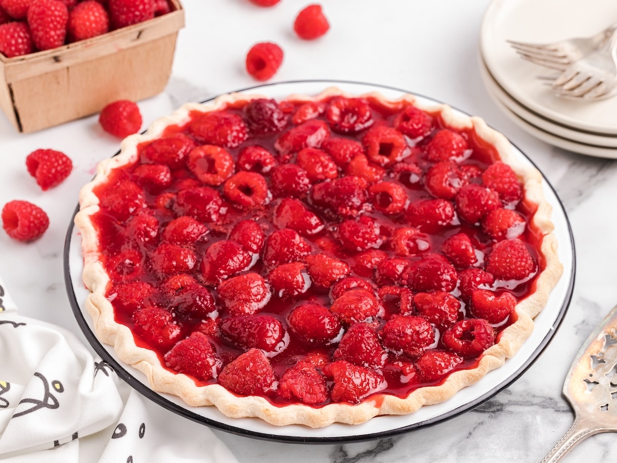 A fresh raspberry pie filled with glossy raspberry filling and whole raspberries sits in a flaky pie crust on a marble countertop, surrounded by scattered raspberries, stacked dessert plates, and a basket of fresh berries.