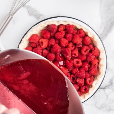 Bright red raspberry glaze is being poured from a saucepan over fresh raspberries in an unbaked pie crust, with a whisk resting nearby on a white marble countertop.