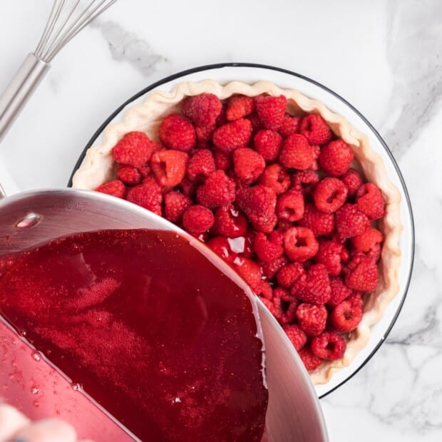Bright red raspberry glaze is being poured from a saucepan over fresh raspberries in an unbaked pie crust, with a whisk resting nearby on a white marble countertop.