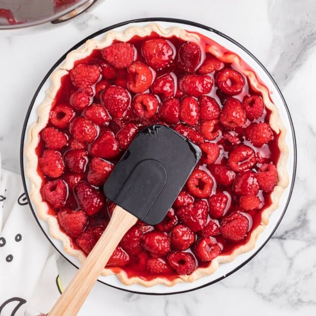 A raspberry pie filling made with whole raspberries and strawberries sits inside an unbaked pie crust, with glossy red filling being spread evenly using a black silicone spatula on a white marble countertop.