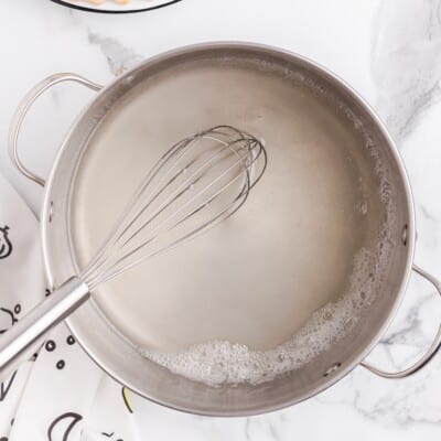 A stainless steel saucepan filled with a cloudy white thickened sauce sits on a white marble countertop, with a metal whisk resting inside the pot and light foam forming around the edges of the liquid.