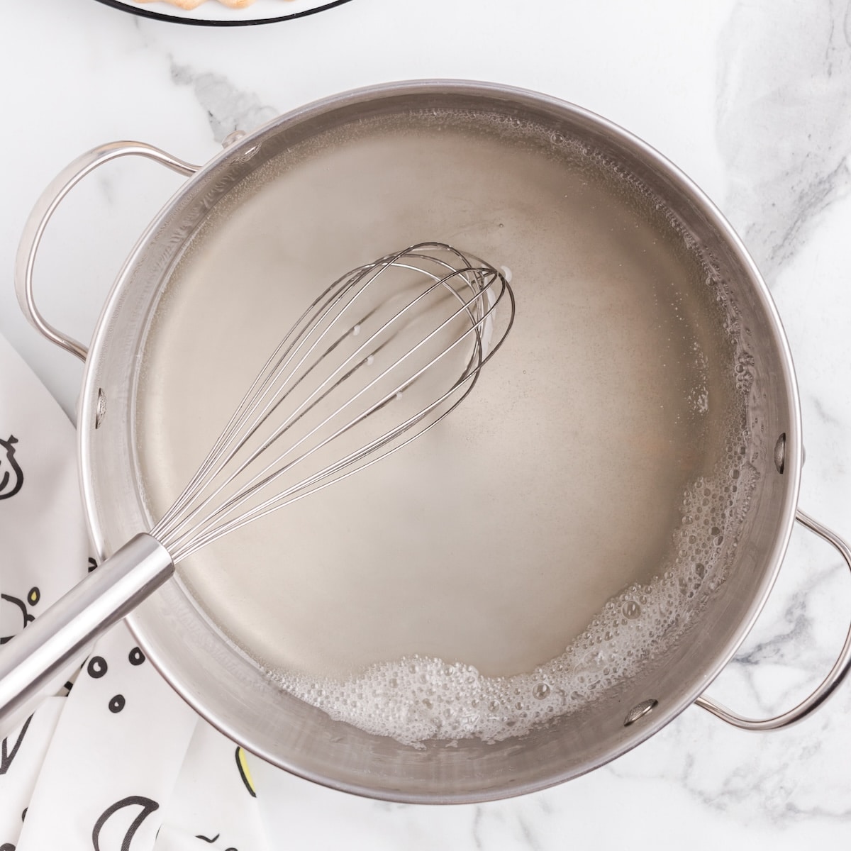A stainless steel saucepan filled with a cloudy white thickened sauce sits on a white marble countertop, with a metal whisk resting inside the pot and light foam forming around the edges of the liquid.