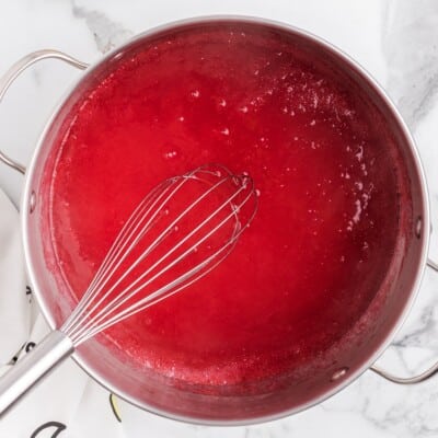 A stainless steel saucepan filled with bright red raspberry pie filling simmers on a white marble countertop, with small bubbles forming on the surface and a metal whisk resting inside the pot.