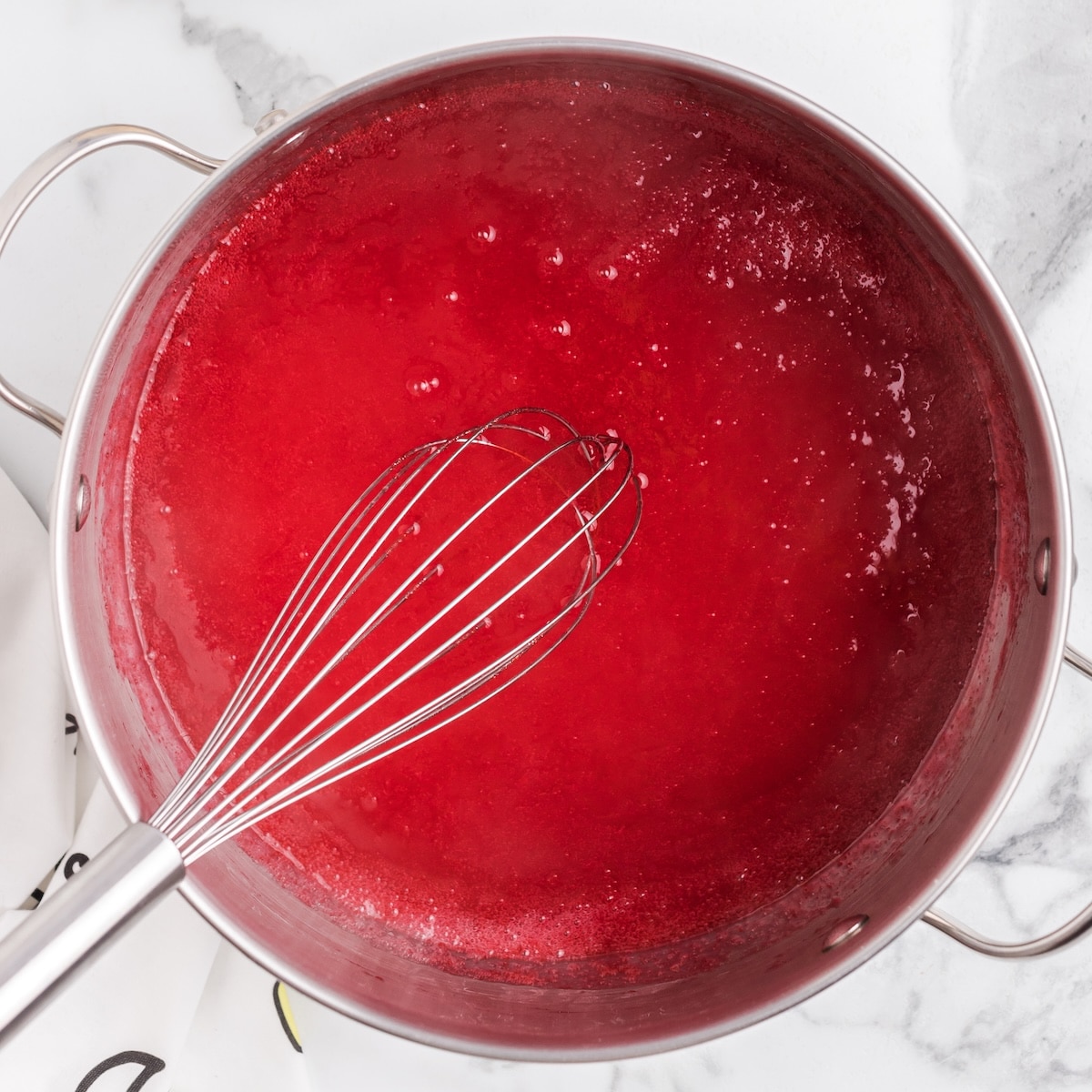 A stainless steel saucepan filled with bright red raspberry pie filling simmers on a white marble countertop, with small bubbles forming on the surface and a metal whisk resting inside the pot.