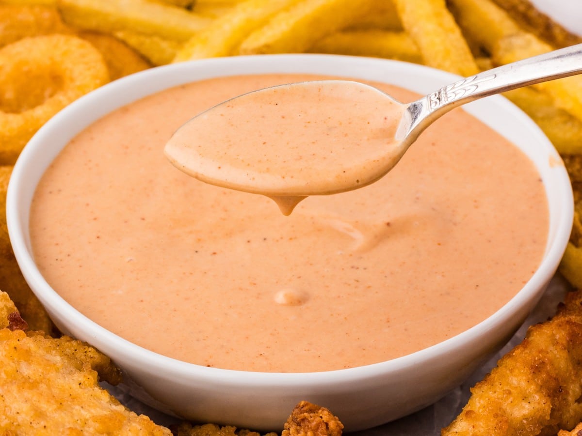 Creamy Red Robin Campfire Sauce in a white serving bowl with a spoon lifting the smoky dipping sauce, surrounded by crispy french fries and golden fried chicken tenders.