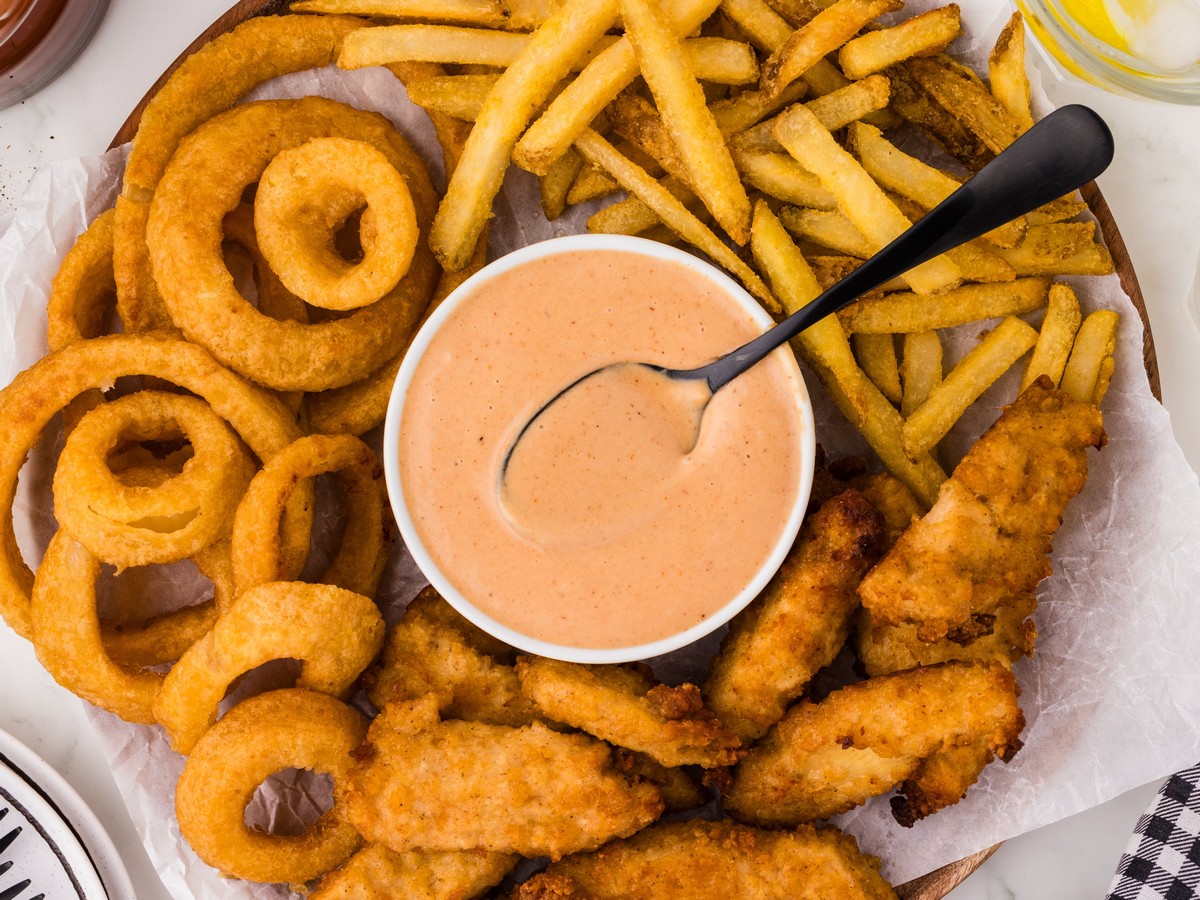 Overhead view of Red Robin Campfire Sauce served in a white bowl with a spoon, surrounded by crispy chicken tenders, onion rings, and seasoned french fries on a serving platter.