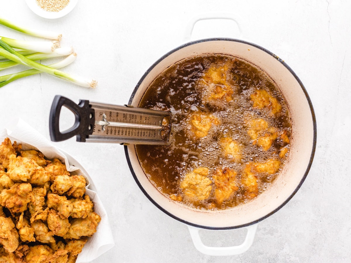 Breaded chicken pieces frying in a Dutch oven with a clip-on thermometer monitoring the oil temperature, with a batch of cooked crispy chicken draining nearby.