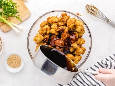 Pouring homemade sesame sauce over crispy fried chicken pieces in a large glass bowl, with sliced green onions and toasted sesame seeds nearby.