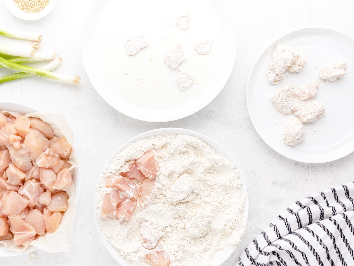 Diced chicken breast being coated in seasoned flour as part of the buttermilk double-dredge process for crispy sesame chicken.
