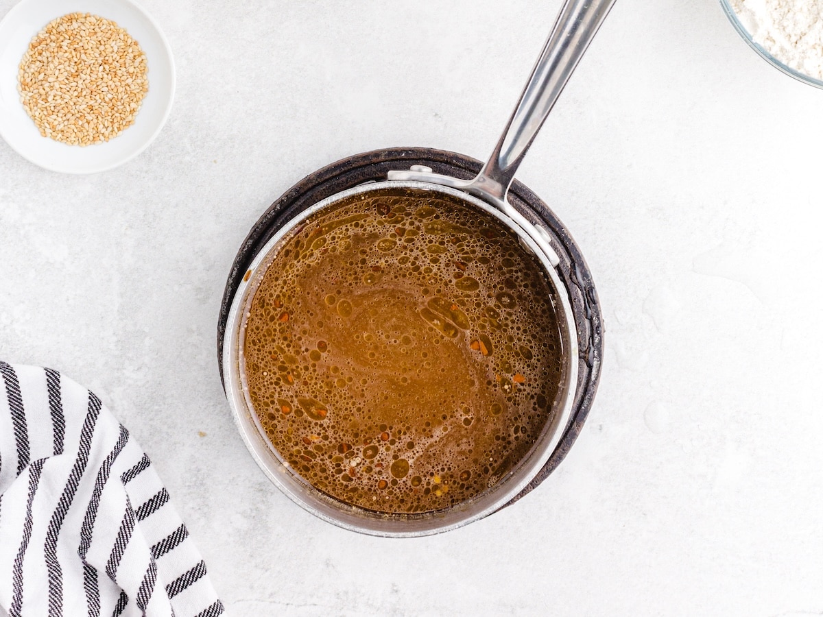 Sesame chicken sauce simmering in a small saucepan with toasted sesame seeds and flour visible in the background.