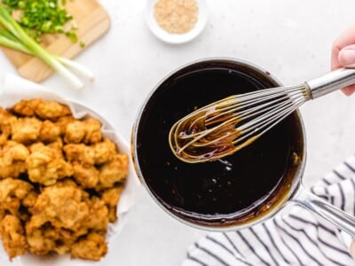 Whisking the homemade sesame sauce in a saucepan next to a bowl of crispy fried chicken pieces ready to be tossed.